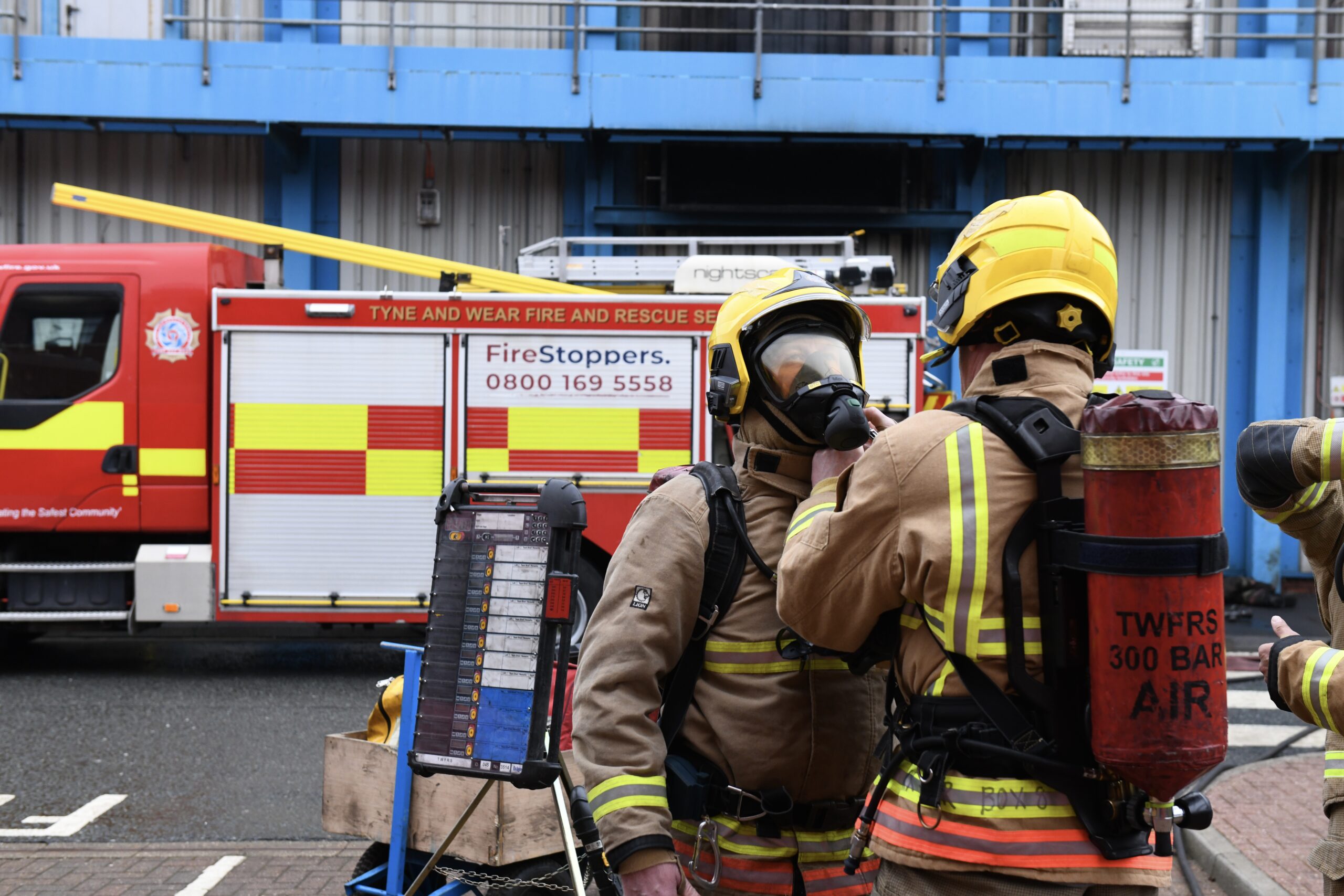 Two firefighters, wearing full gear including breathing apparatus, standing outside a fire engine