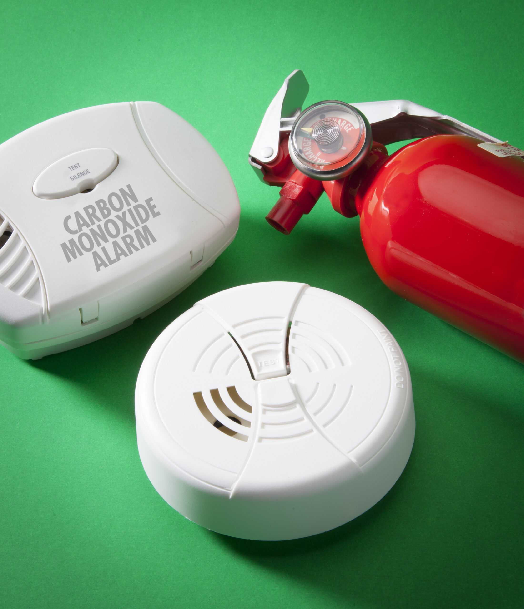 A photograph of three pieces of fire safety equipment on a green background from left to right a carbon monoxide alarm, a smoke alarm and a red fire extinguisher.