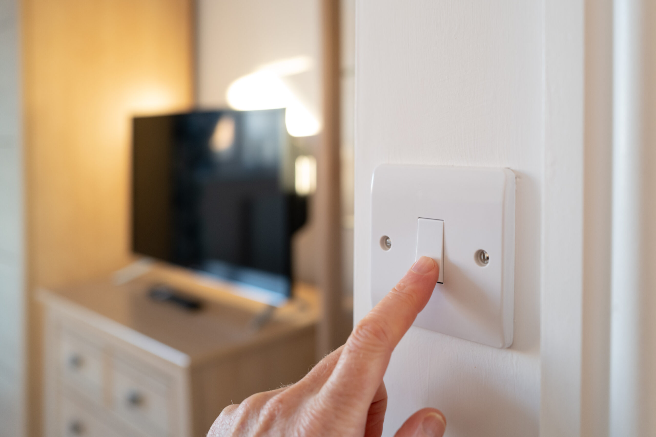 A photograph of a finger pressing a white light switch with a blurred television on a chest of drawers in the background.