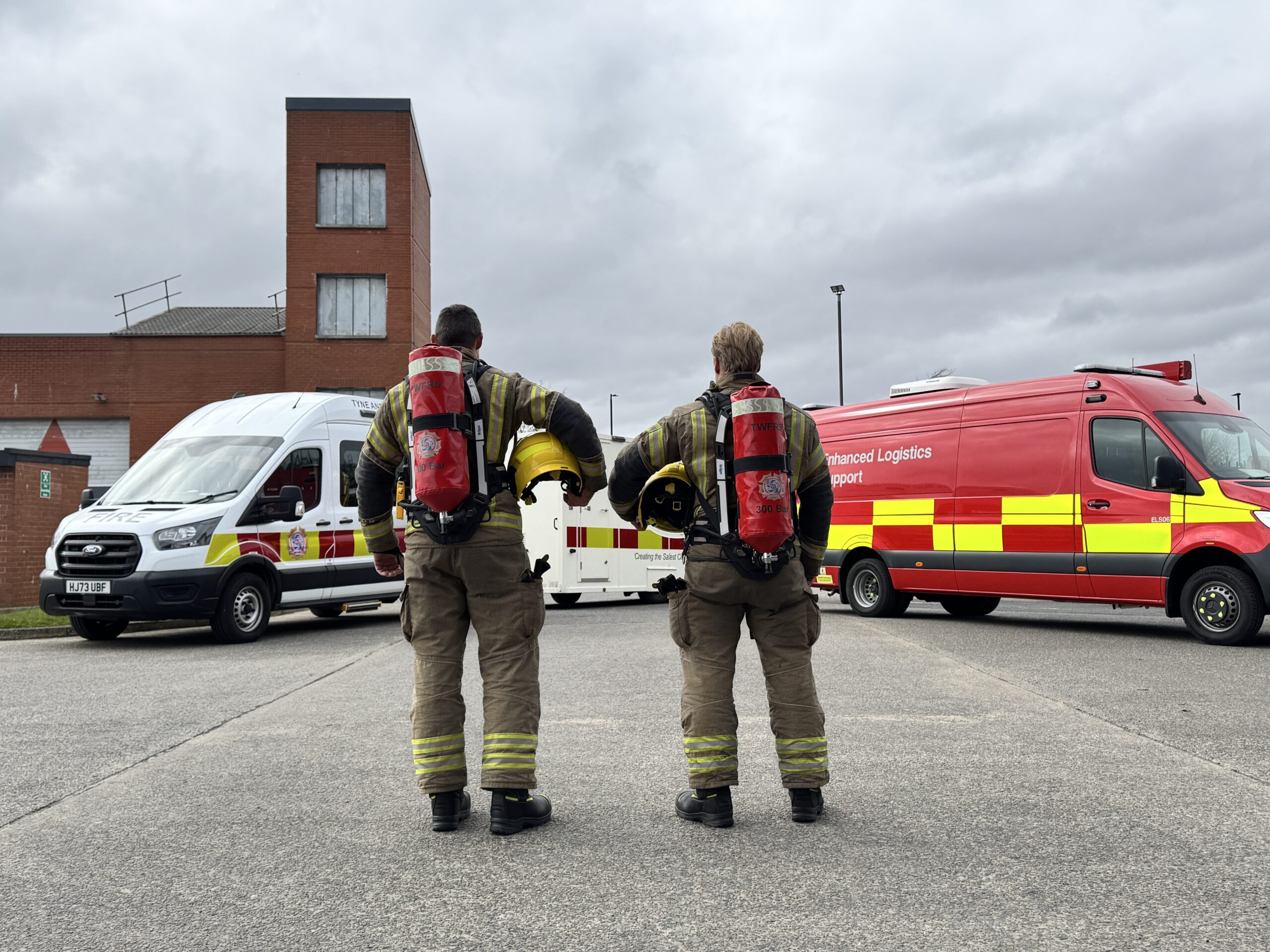 Photograph of two firefighters wearing fire kit, breathing apparatus, standing in front of the welfare vehicle and enhanced logistic support unit