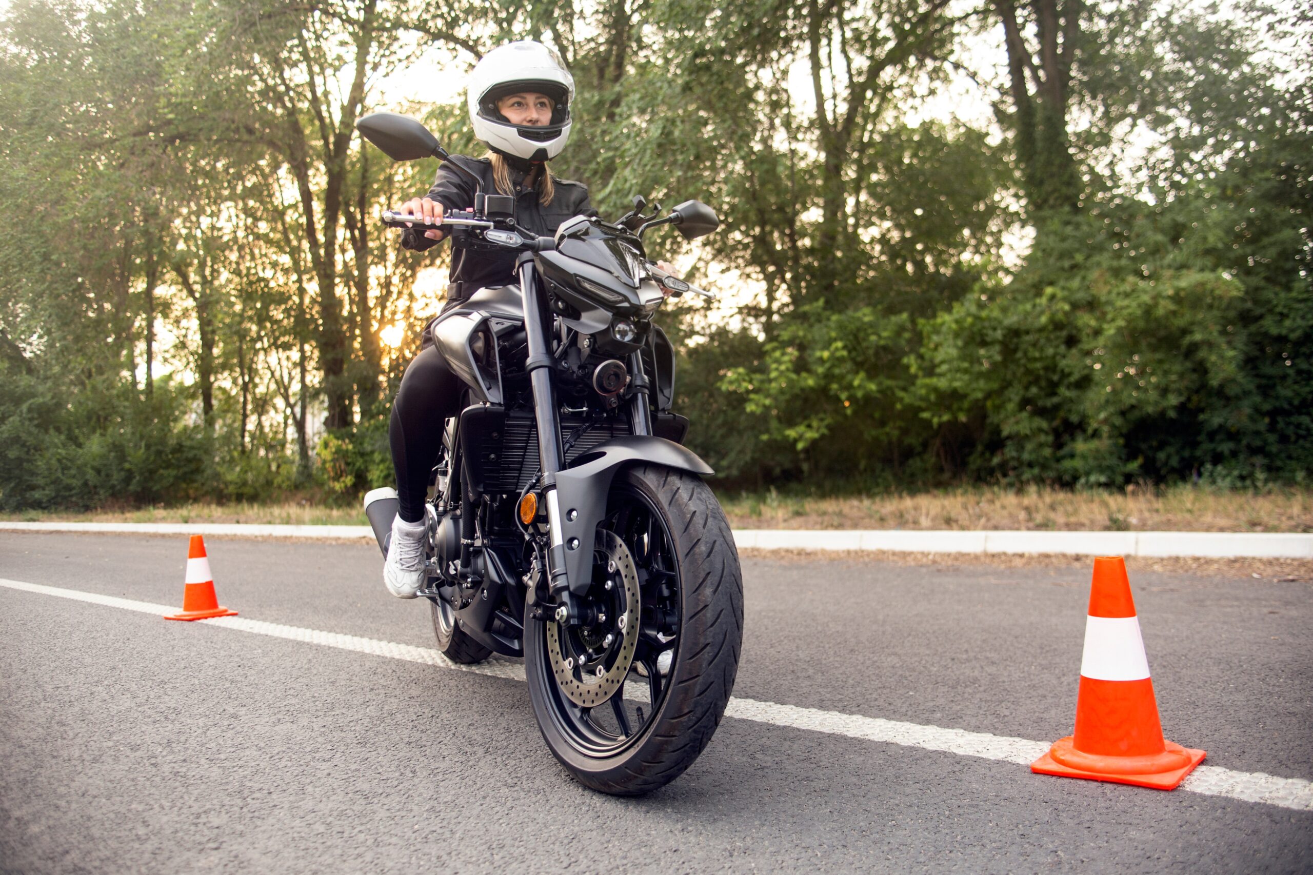 A female motorcyclist sitting on a black motorbike positioned between two orange and white bollards with trees in the background.