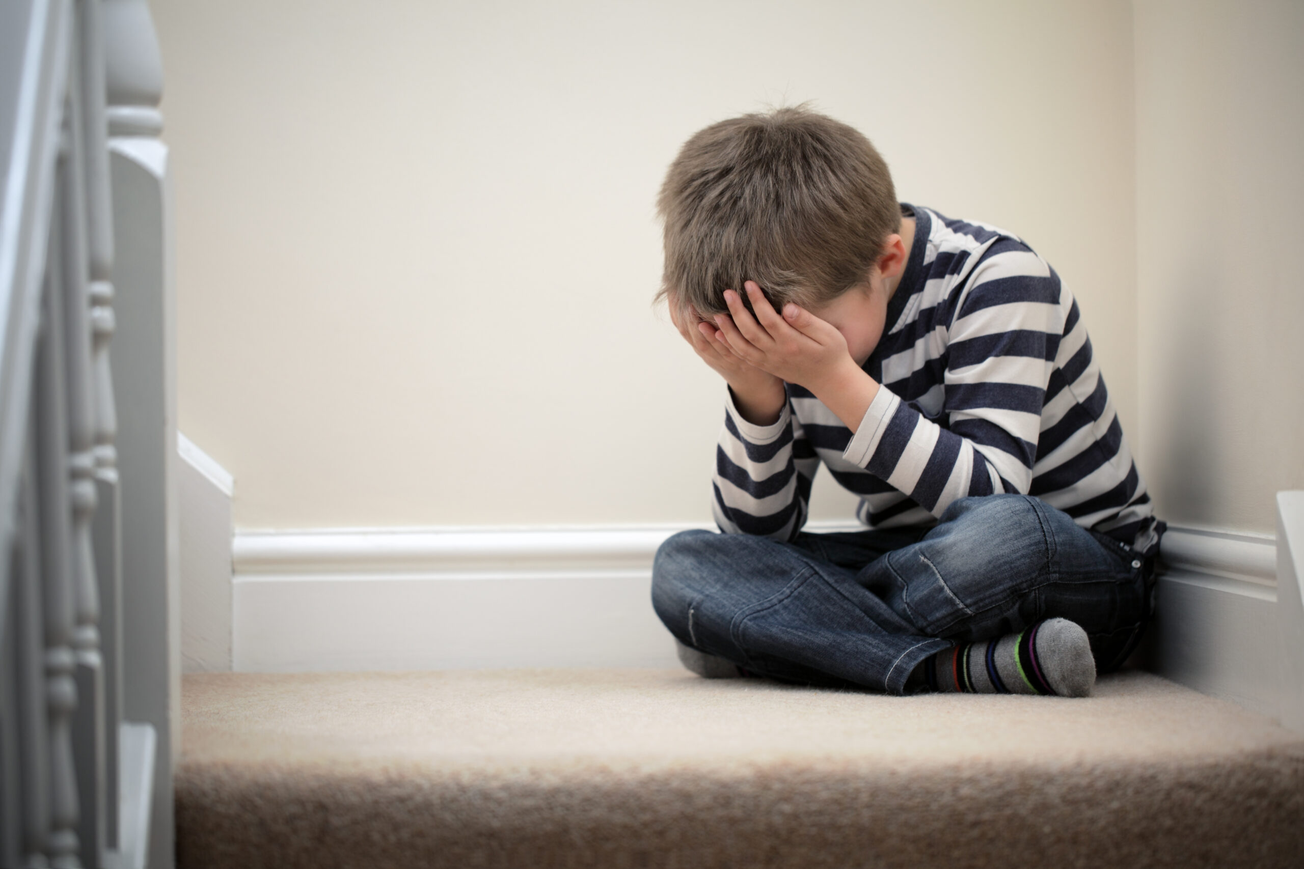 Safeguarding - a young boy wearing a stripy black and white tshirt with his head in his hands covering his face. Is sat cross legged at the top of a carpeted stairs.