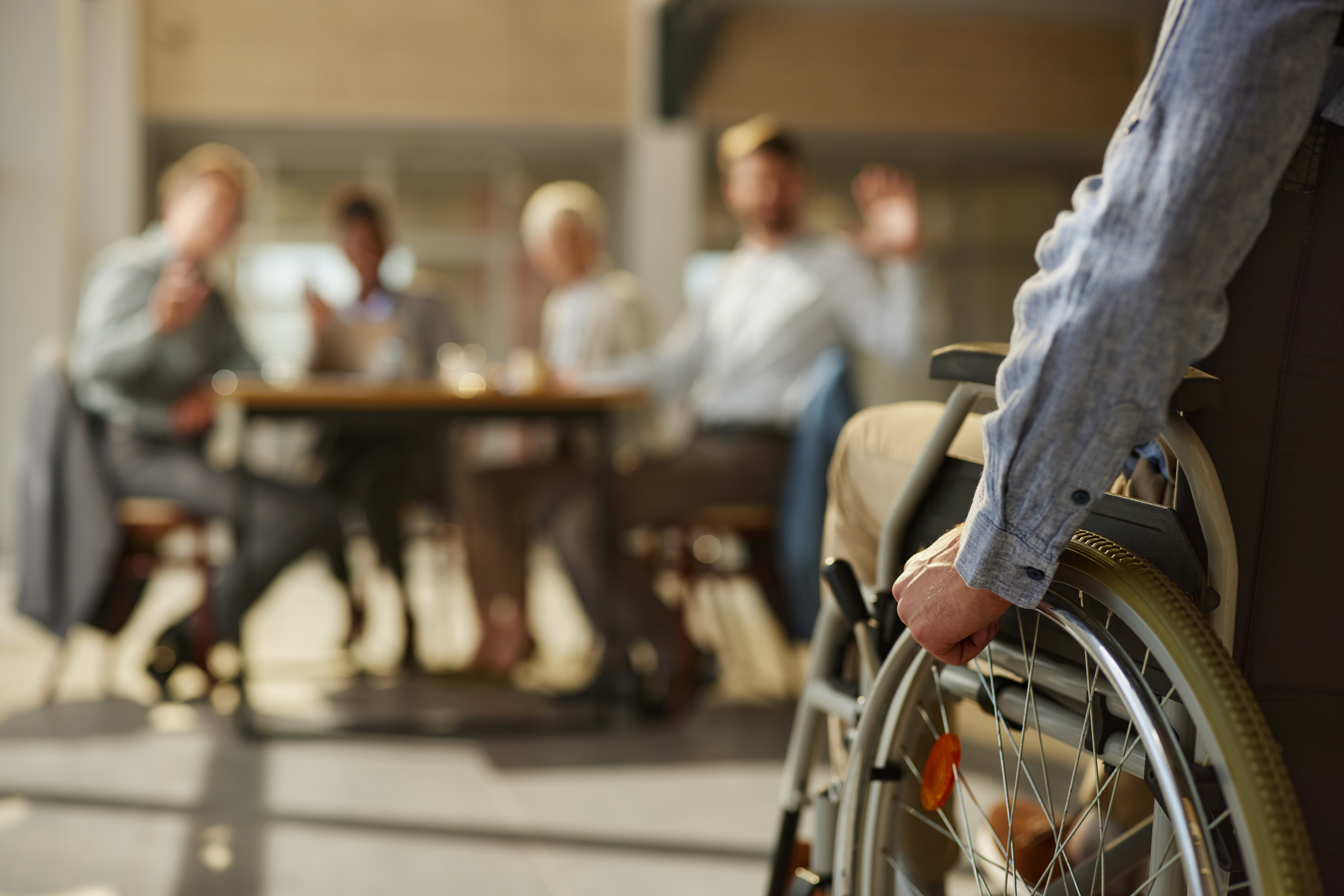 A photograph of a person in a wheelchair positioned to the right of the picture with the arm and side of the wheelchair in view. Background is blurred.