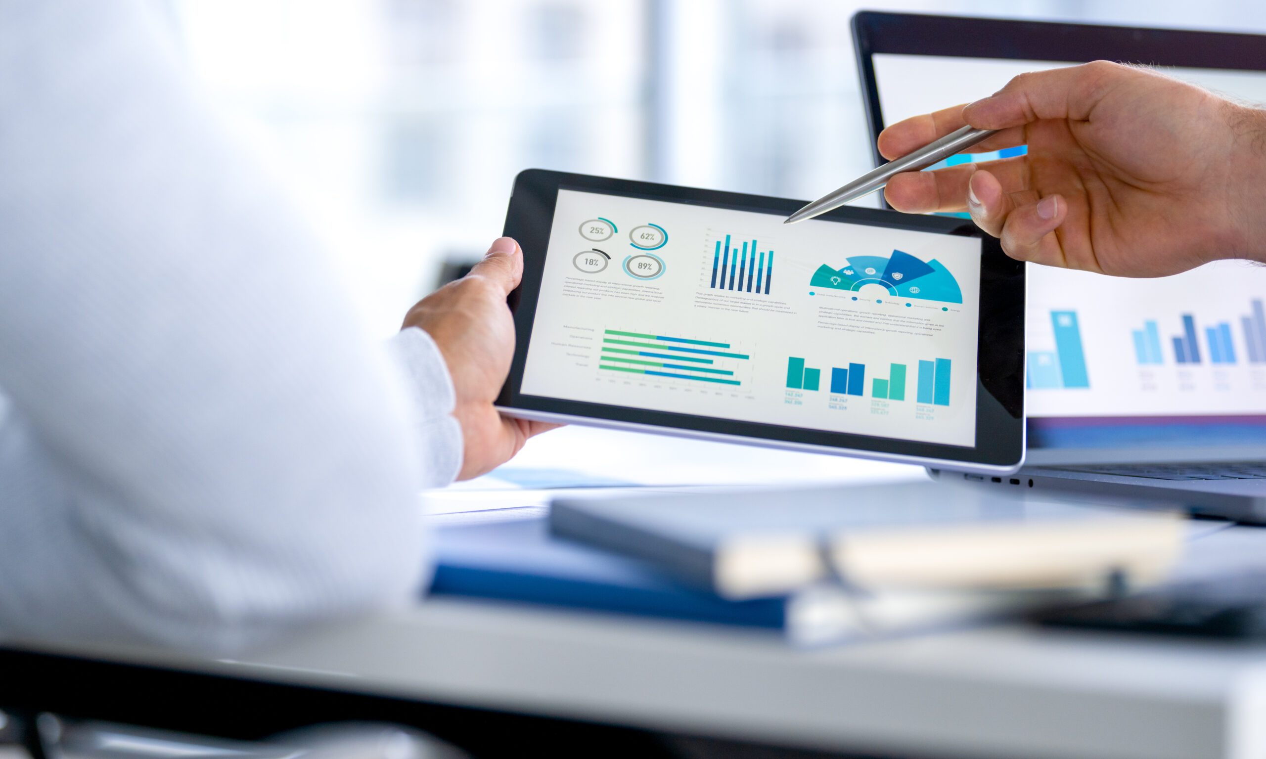 Two business people meeting and looking at financial data, charts and graphs on a digital tablet and a laptop computer. There are documents and paperwork on the table. One man is pointing at the screen.